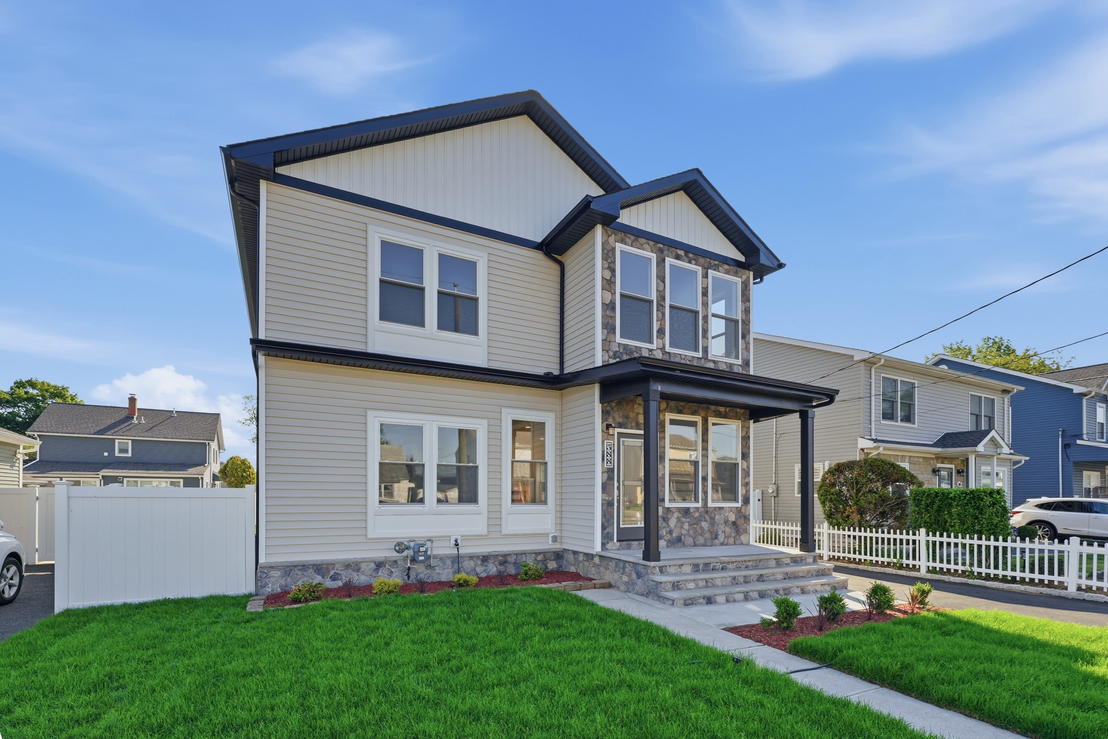 Modern two-story home with tan siding, stone accents, black trim, and a manicured green lawn.