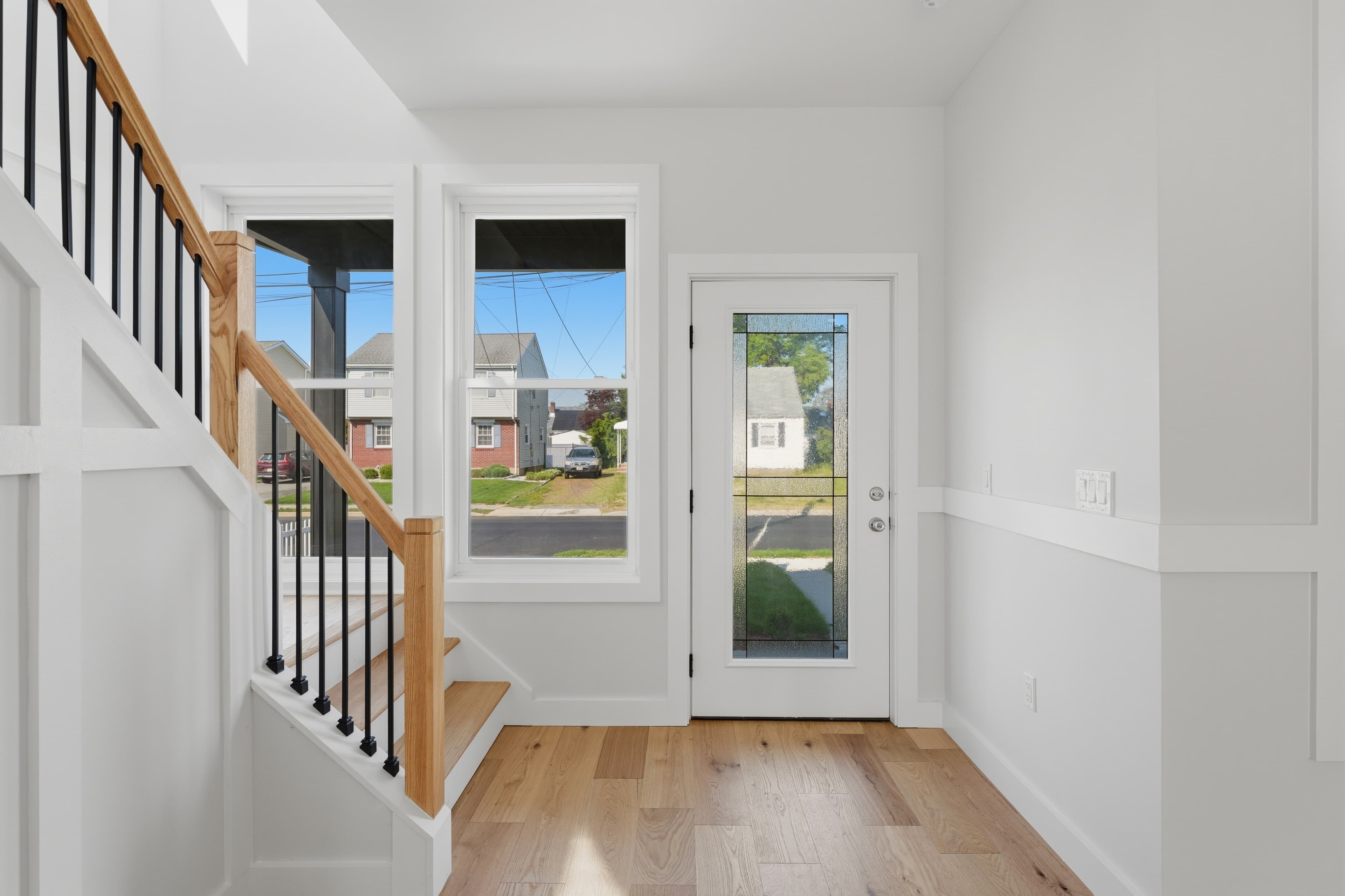 Modern white foyer featuring light wood floors, a wooden staircase, and large bright windows.