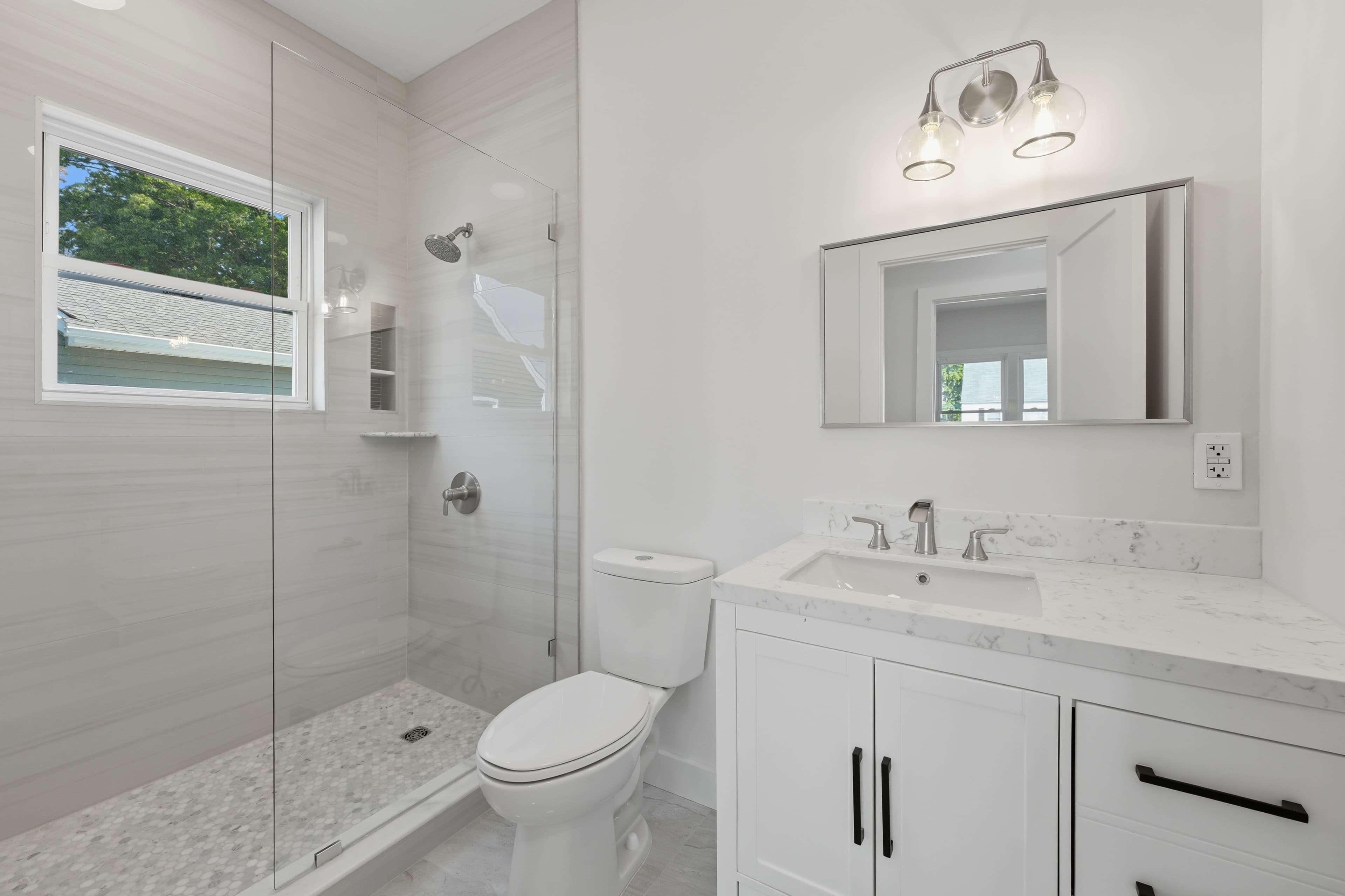 Modern white bathroom with a glass shower, marble vanity, rectangular mirror, and three-bulb light fixture.