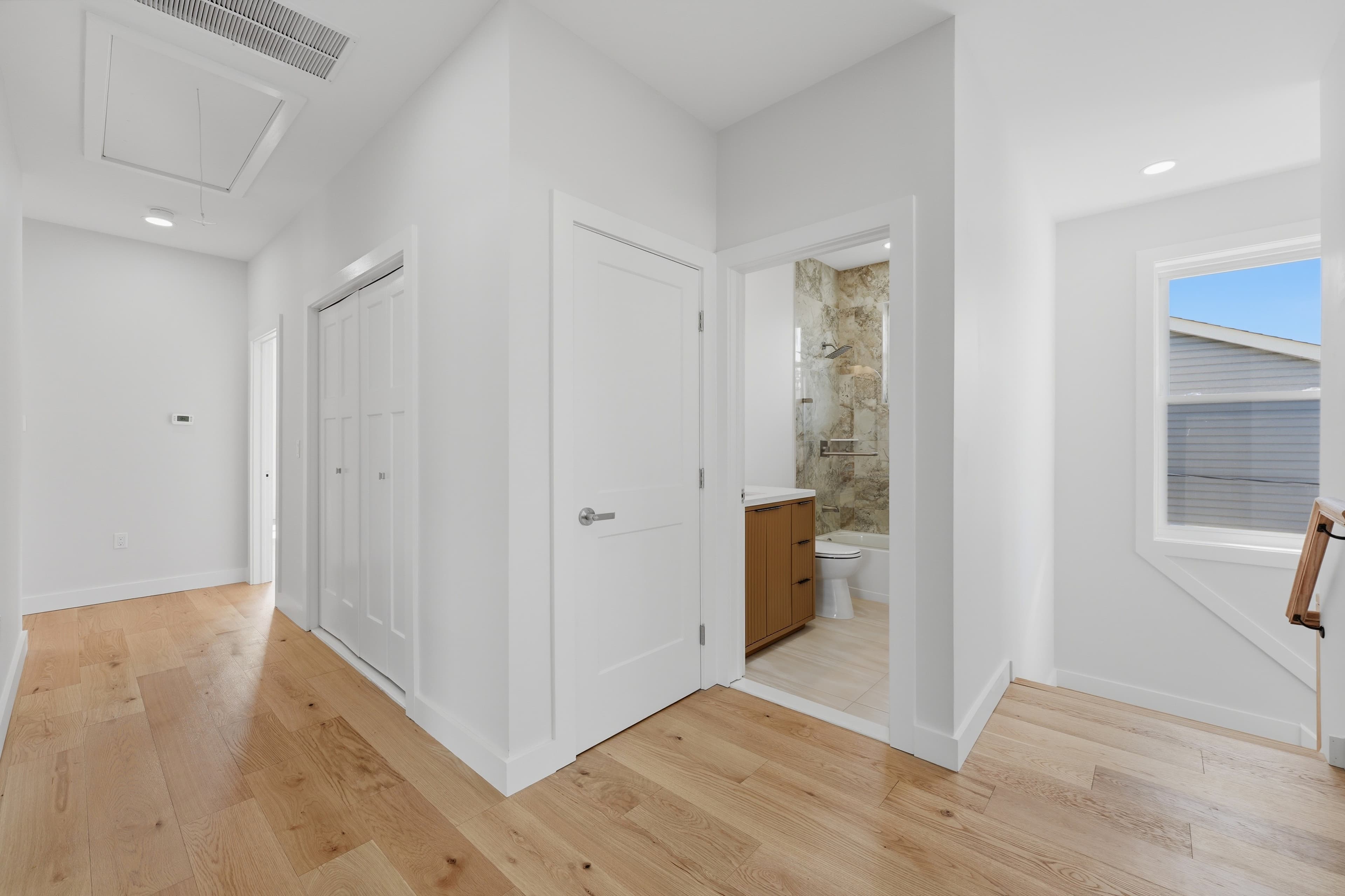 Modern hallway featuring light wood floors, white walls, and an open bathroom doorway.