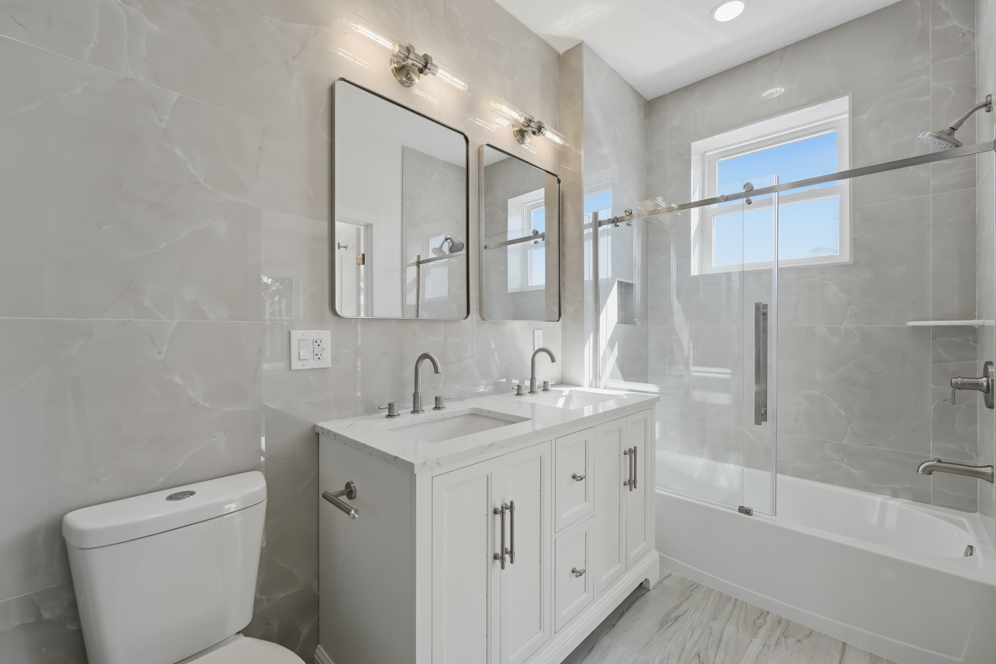 Modern bathroom with white double vanity, twin mirrors, and glass-enclosed bathtub with grey marble walls.