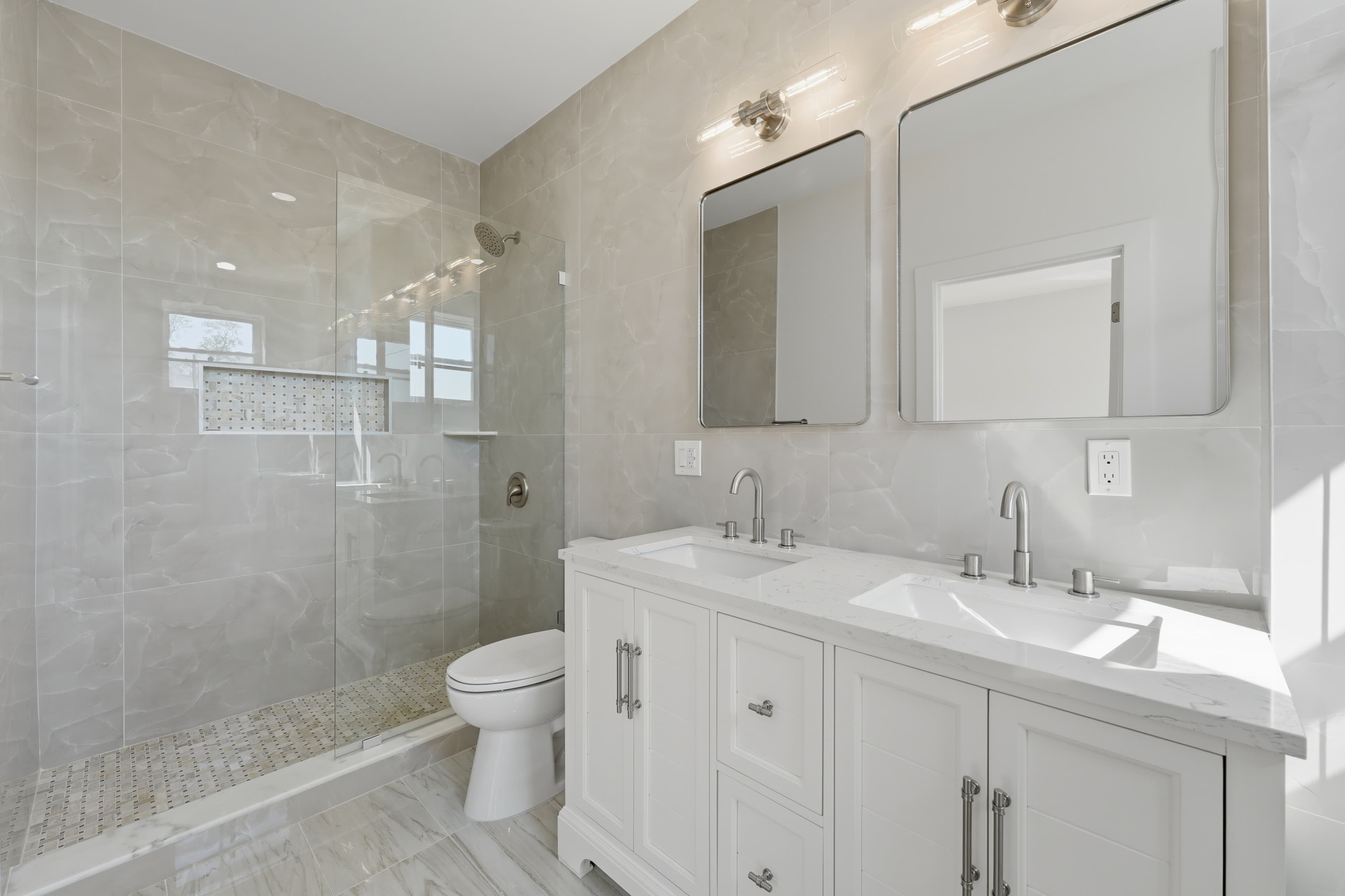 Modern bathroom with a white double vanity, glass-enclosed shower, and beige marble-patterned wall tiles.