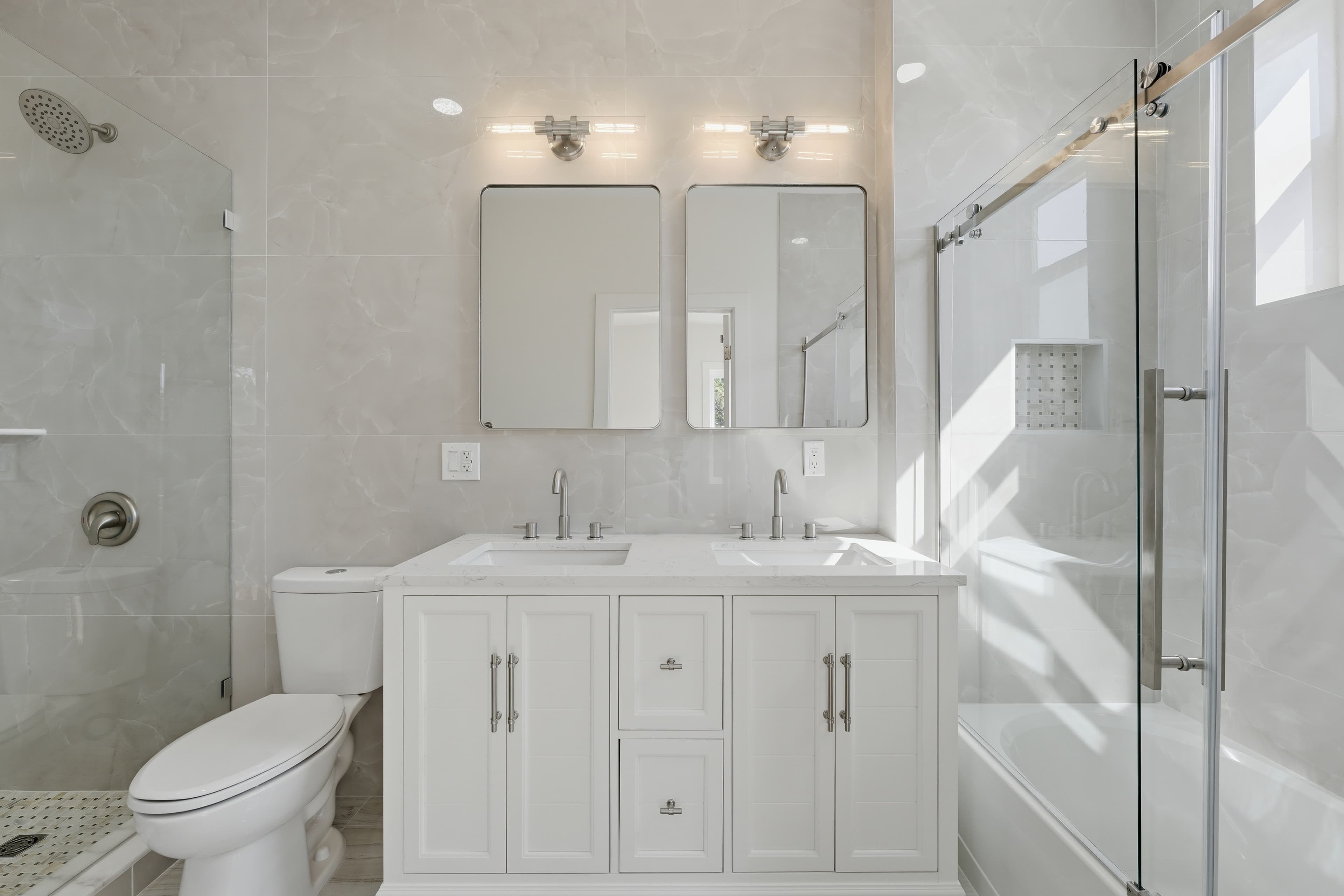 Modern bathroom with a white double vanity, two mirrors, marble tiles, and a glass shower.