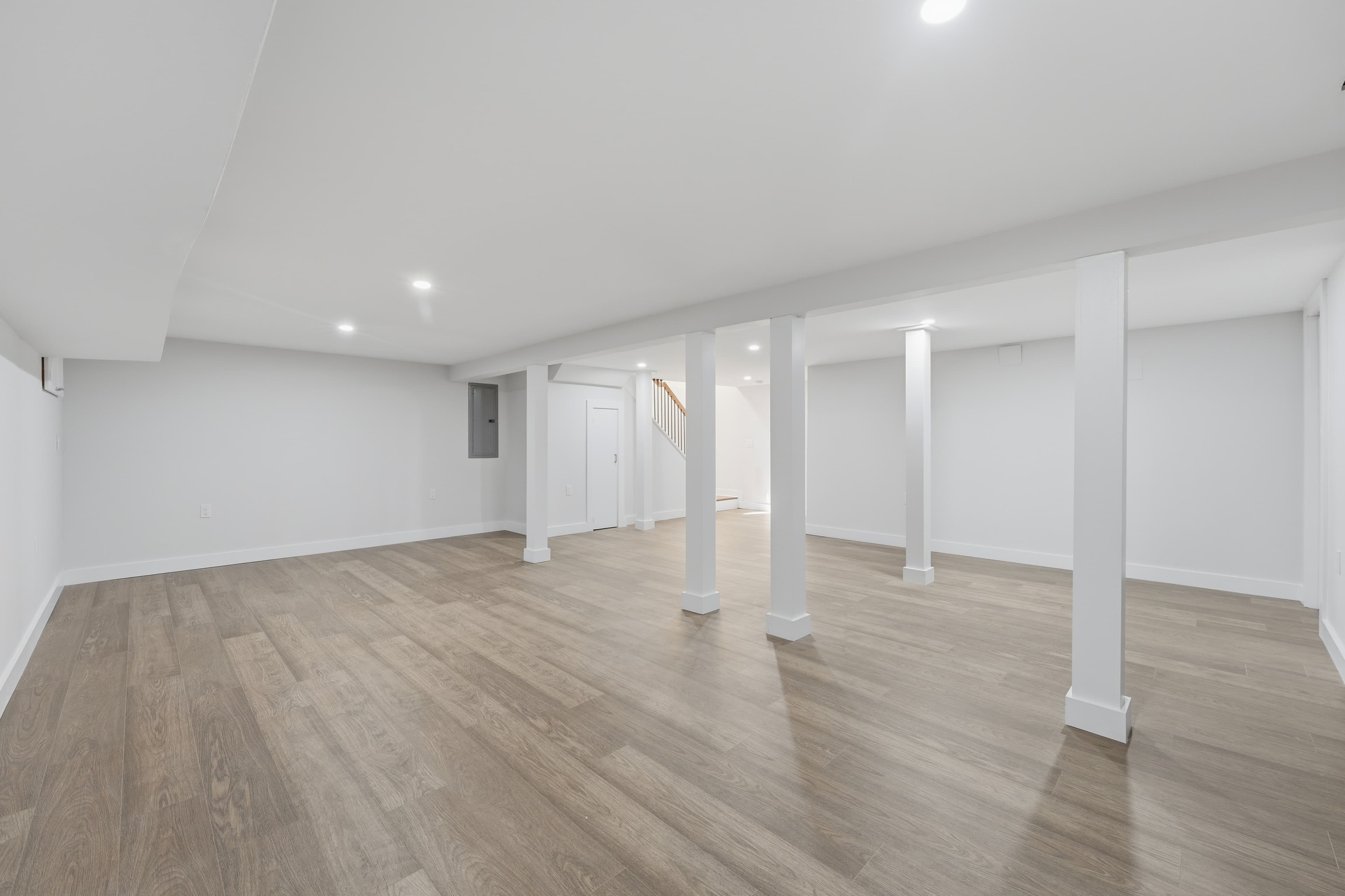 Empty finished basement with white walls, support pillars, recessed lighting, and light wood-look flooring.