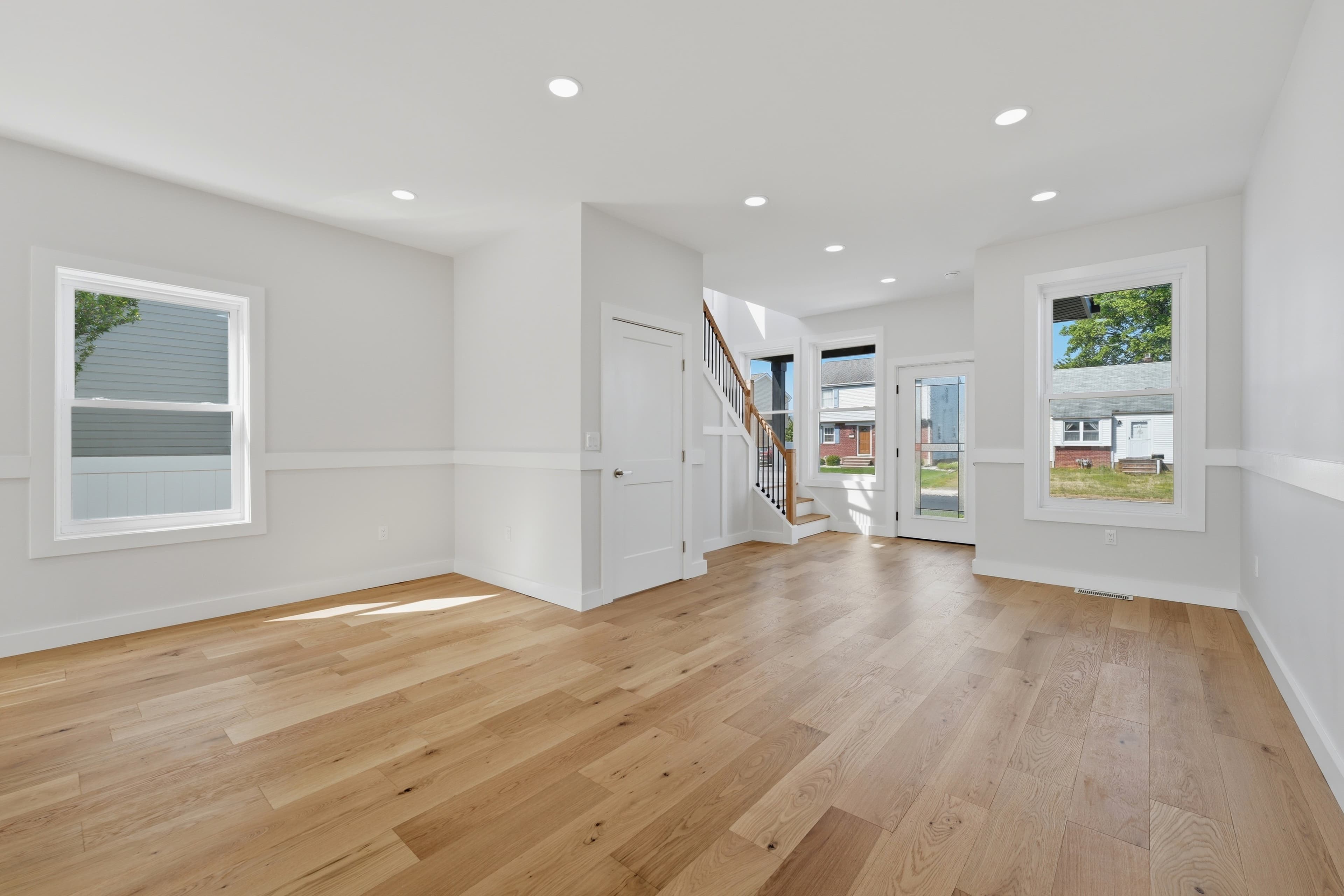 Sunlit empty room with light hardwood floors, white walls, large windows, and a staircase.